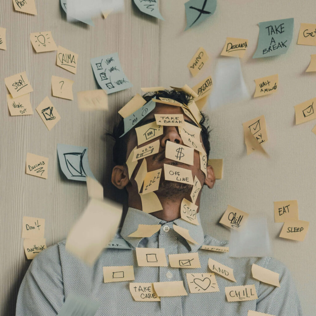 A person is leaning against a wall, overwhelmed with numerous sticky notes covering their face and upper body. The sticky notes contain various handwritten messages and doodles, such as "TAKE A BREAK," "CHILL," "STOP," and "EAT SLEEP." Additional sticky notes with similar messages are scattered around, some floating in mid-air, creating a chaotic and cluttered visual that represents cognitive overload. The person's eyes are closed, suggesting a state of mental exhaustion.