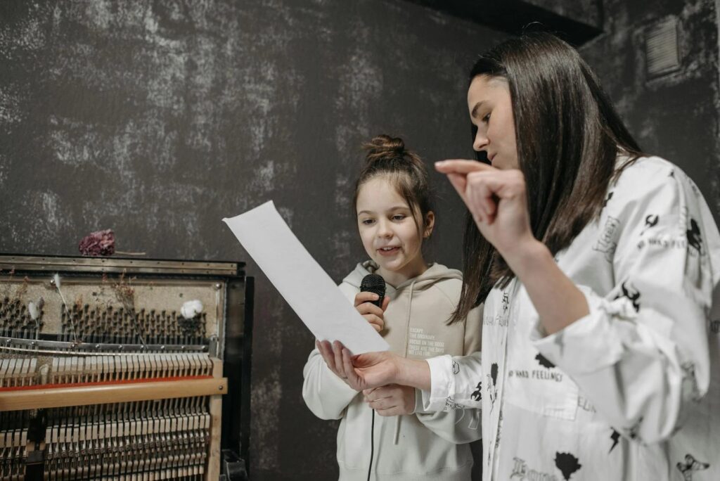 A young girl holding a microphone participates in a collaborative learning activity with an adult who is holding a sheet of paper and gesturing. They are near an upright piano, set against a textured, dark wall, emphasizing student voice in education.