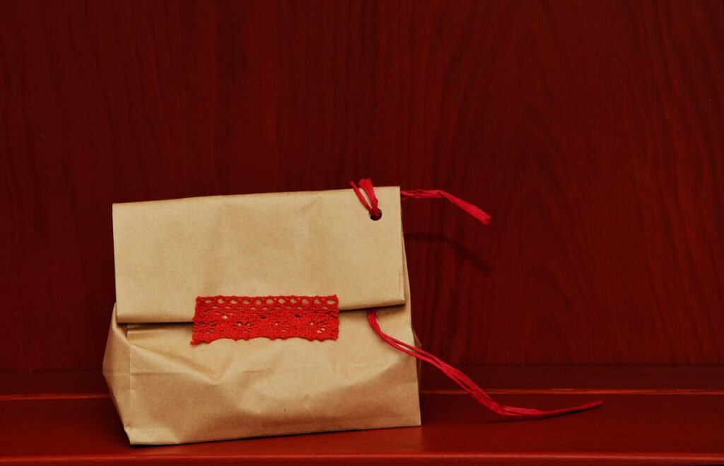 A small brown paper bag is set against a wooden background. The bag is folded at the top and secured with a red ribbon threaded through a punched hole. A strip of red lace decorates the front of the bag, adding a touch of elegance. The setting is simple and rustic, highlighting the handcrafted nature of the gift bag.