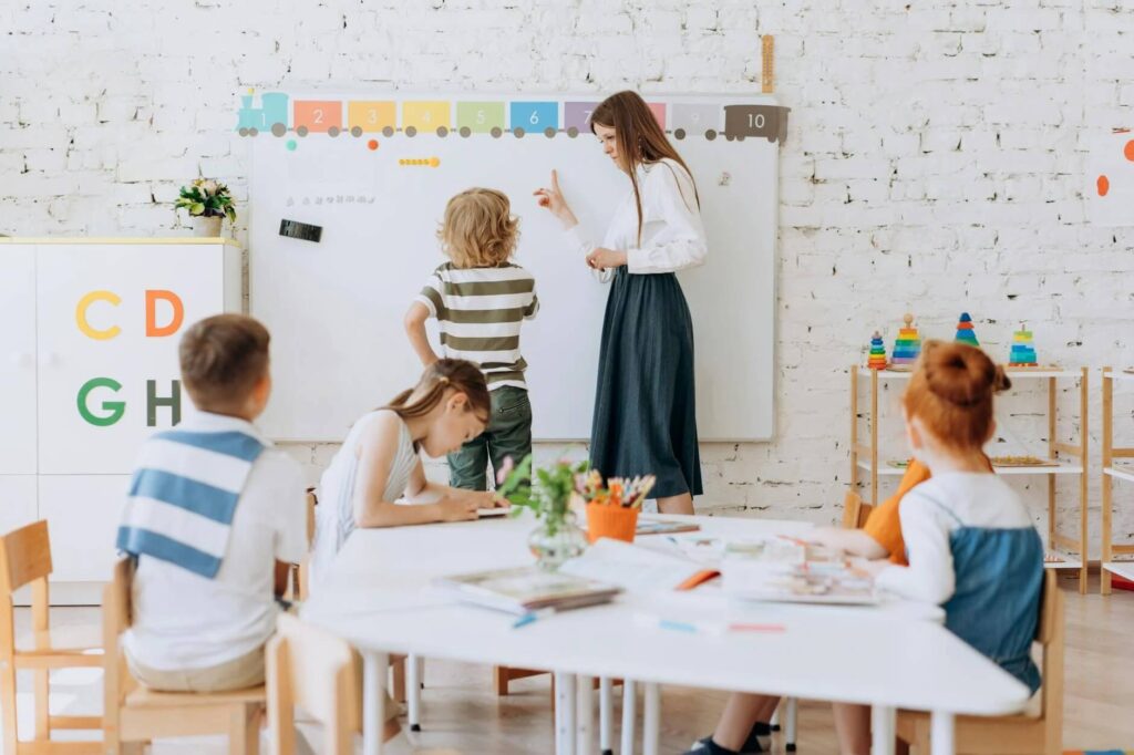A classroom scene with a teacher and young students. The teacher is assisting a child writing on a whiteboard with colorful numbers at the top. Other children are seated around a table, engaged in activities, with colored pencils and papers scattered on the table. The room is bright with educational materials displayed, and plants add a touch of greenery.




